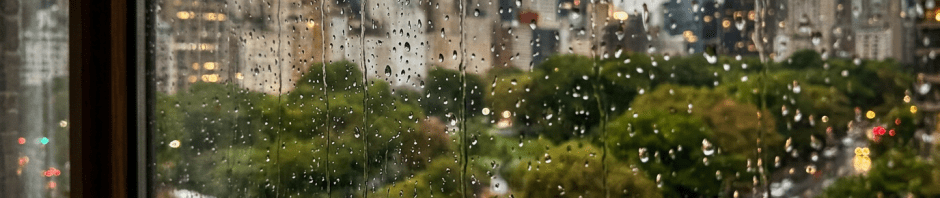 Raindrops on window glass with blurred view of city buildings, trees, and cars on a rainy day